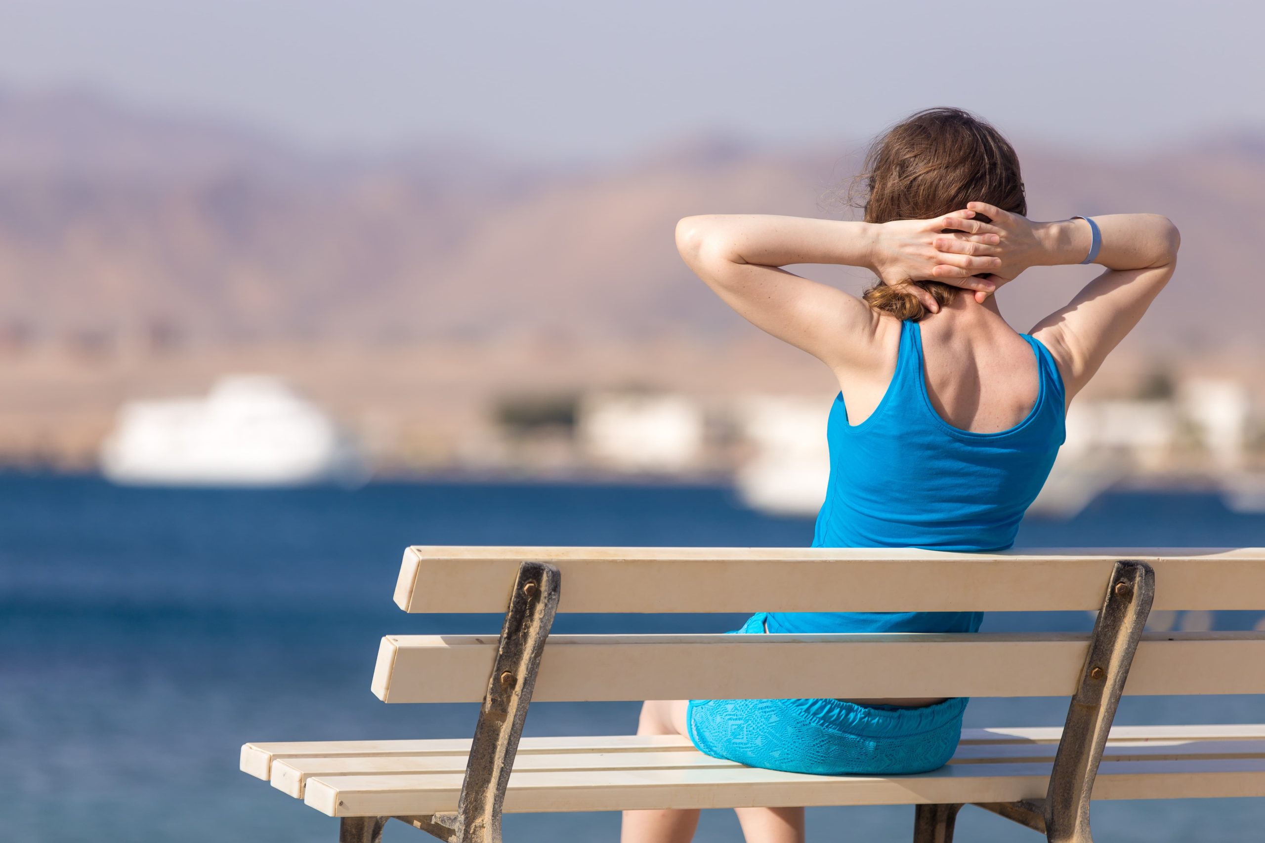 Mujer sentada en un banco en la playa, con las manos detrás de la nuca.